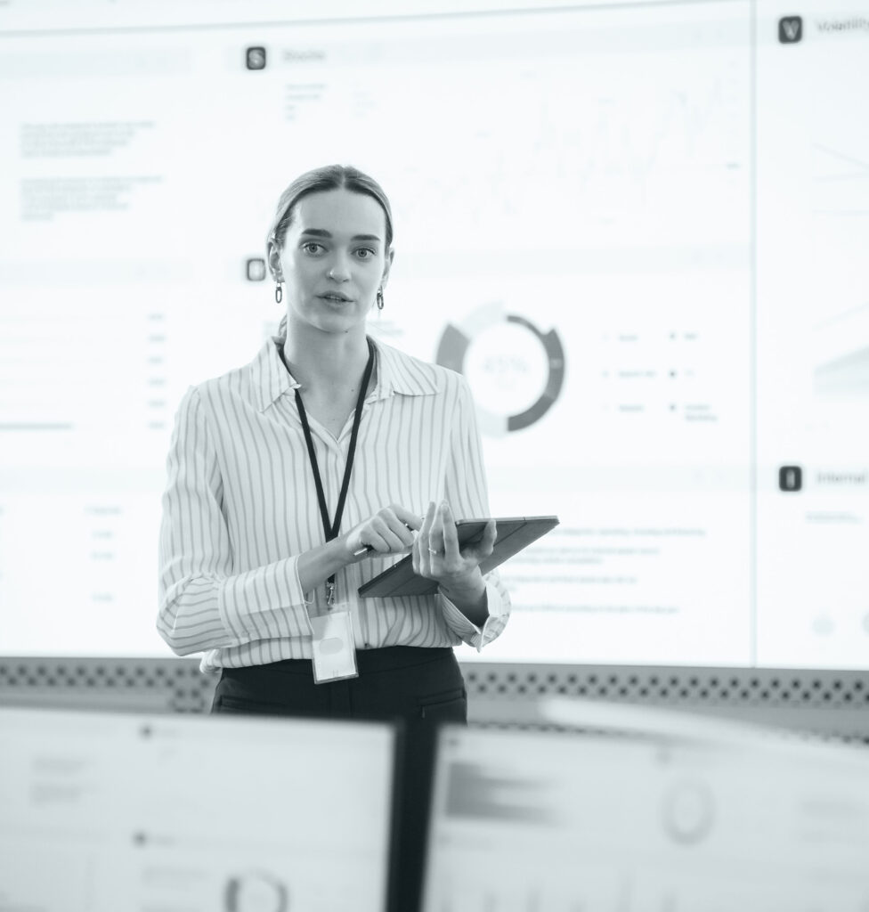 Female Project Manager With Tablet Computer Is Giving Presentation To Diverse Employees In Front of Big Digital Screen In Monitoring Room. Team Of Data Scientists Listening To New Objectives, Tasks.